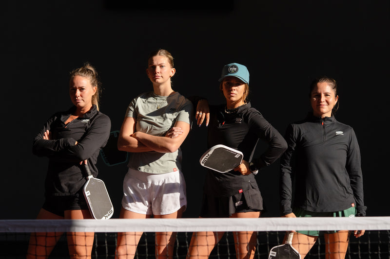 Four women standing behind a net, each holding a paddle, against a black background