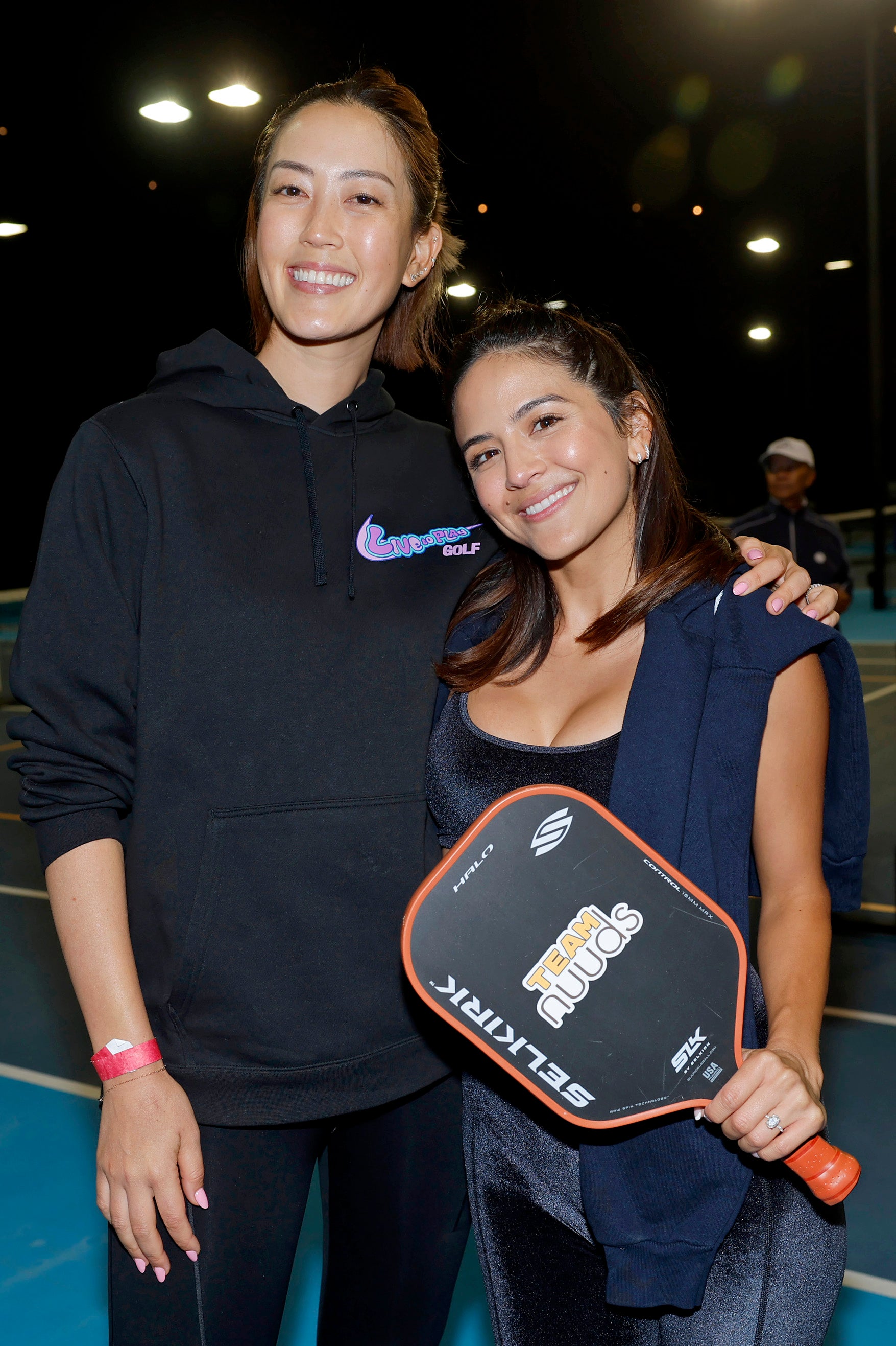 Two women smiling together at a pickleball event, one holding a pickleball paddle.