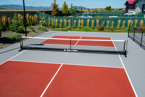 Outdoor sports court with a portable net on brown surface, yellow boundary lines, chain-link fence and trees beyond.