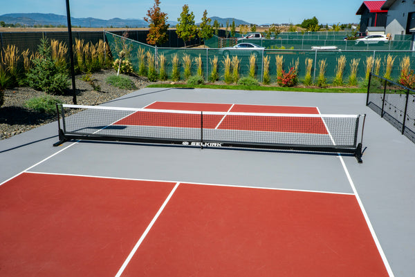 Selkirk Semi-Permanent Pro Pickleball Net set up on a court with red playing surface and green surroundings.