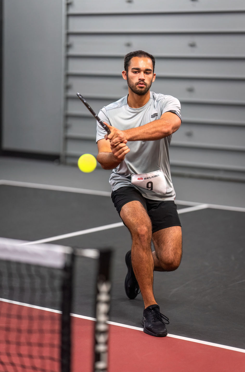 Male pickleball player lunging for a backhand on an indoor court, wearing bib SELKIRK 9