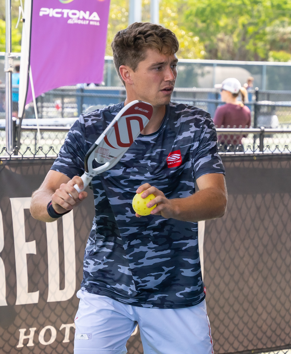 Collin Shick preparing to serve in a pickleball match, wearing a camo shirt.