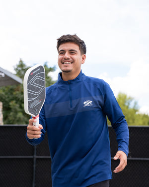 Man smiling while holding a pickleball paddle, wearing a blue Selkirk TruWear Apex 2.0 Quarter Zip Pullover.