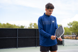 A man wearing a blue Selkirk TruWear Apex 2.0 Quarter Zip Pullover, holding a pickleball paddle on a court.