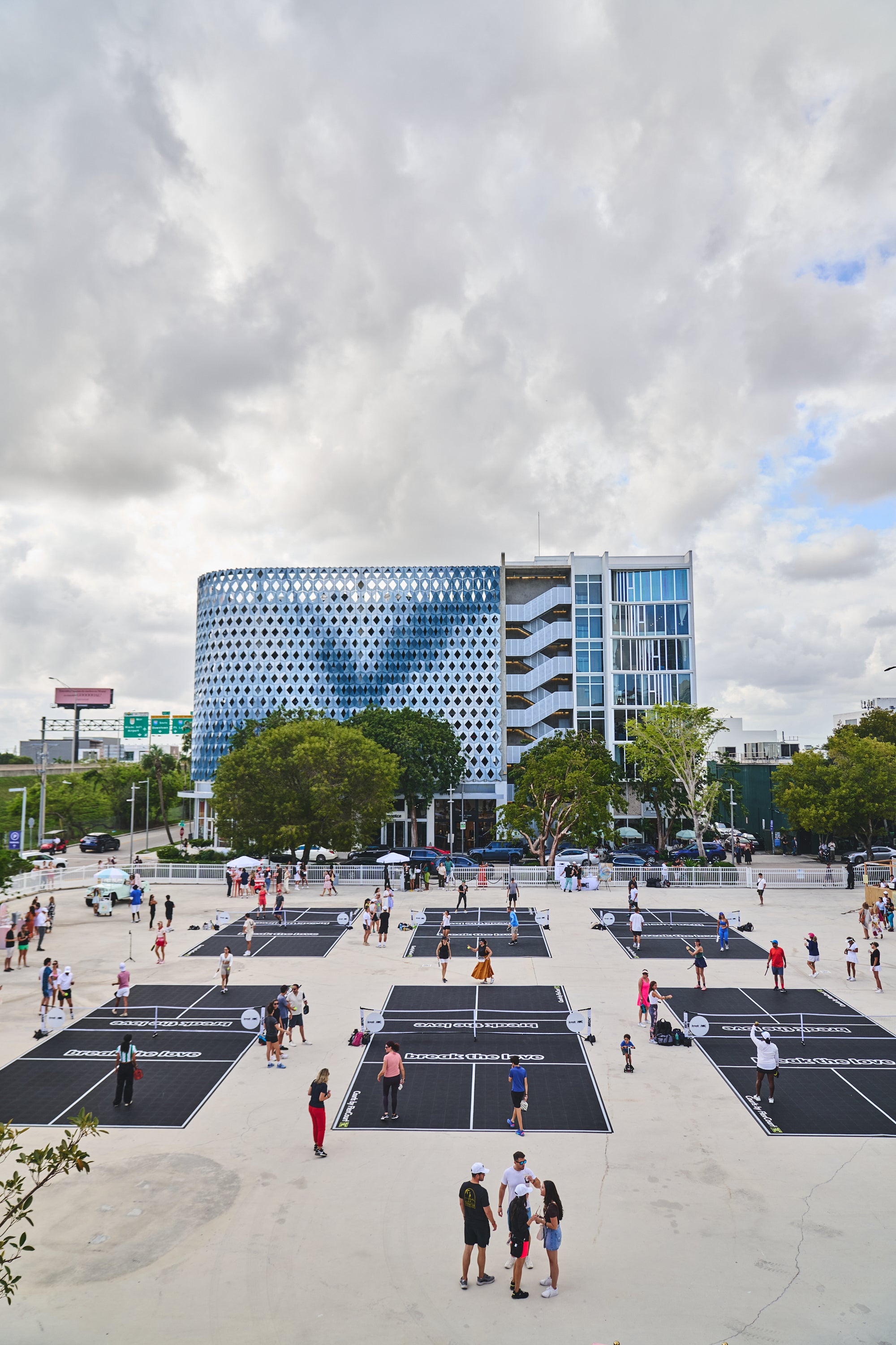 Aerial view of a pickleball event with players on courts and a modern building in the background.