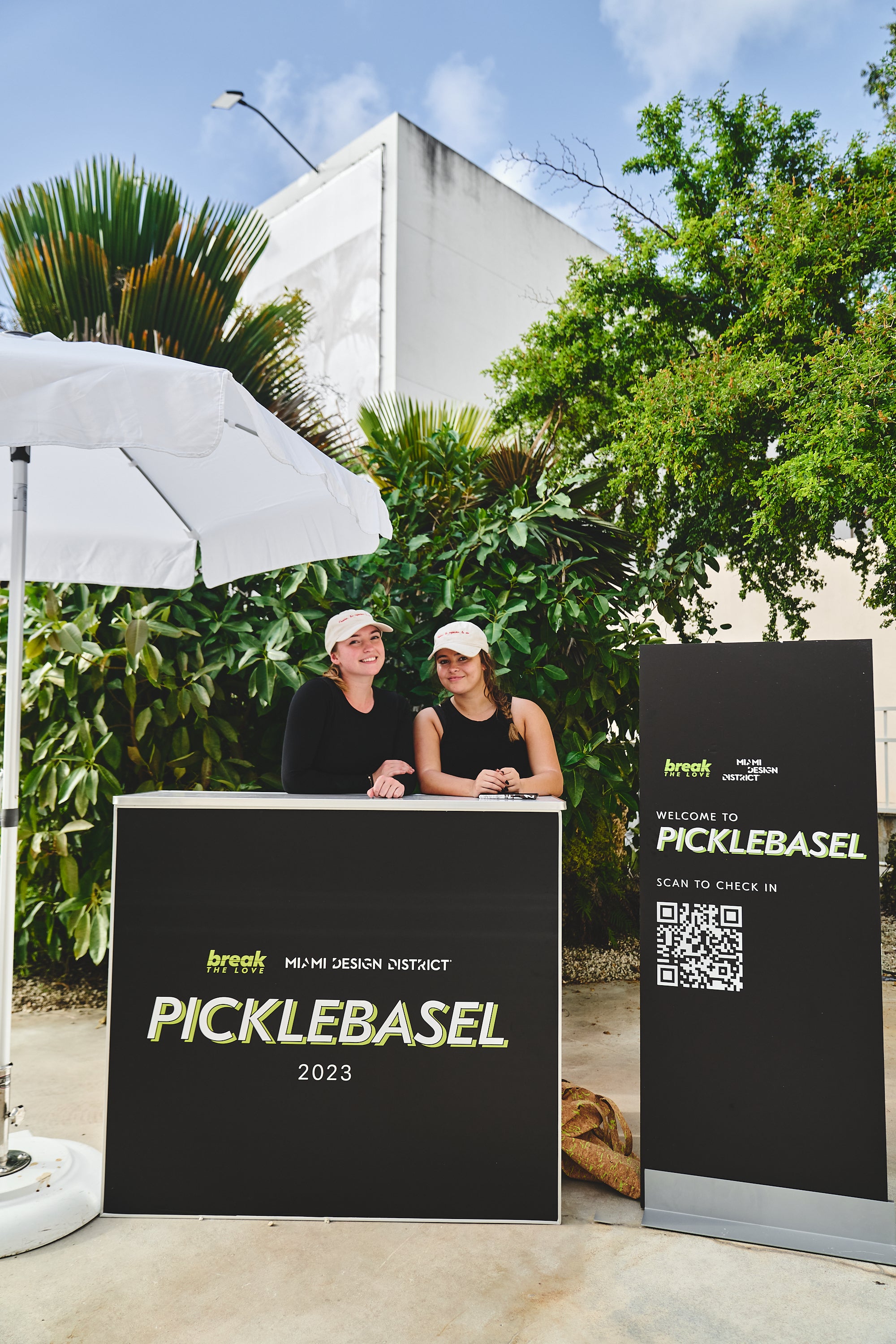 Two women at a welcome booth for PICKLEBASEL 2023, with a QR code and greenery in the background.