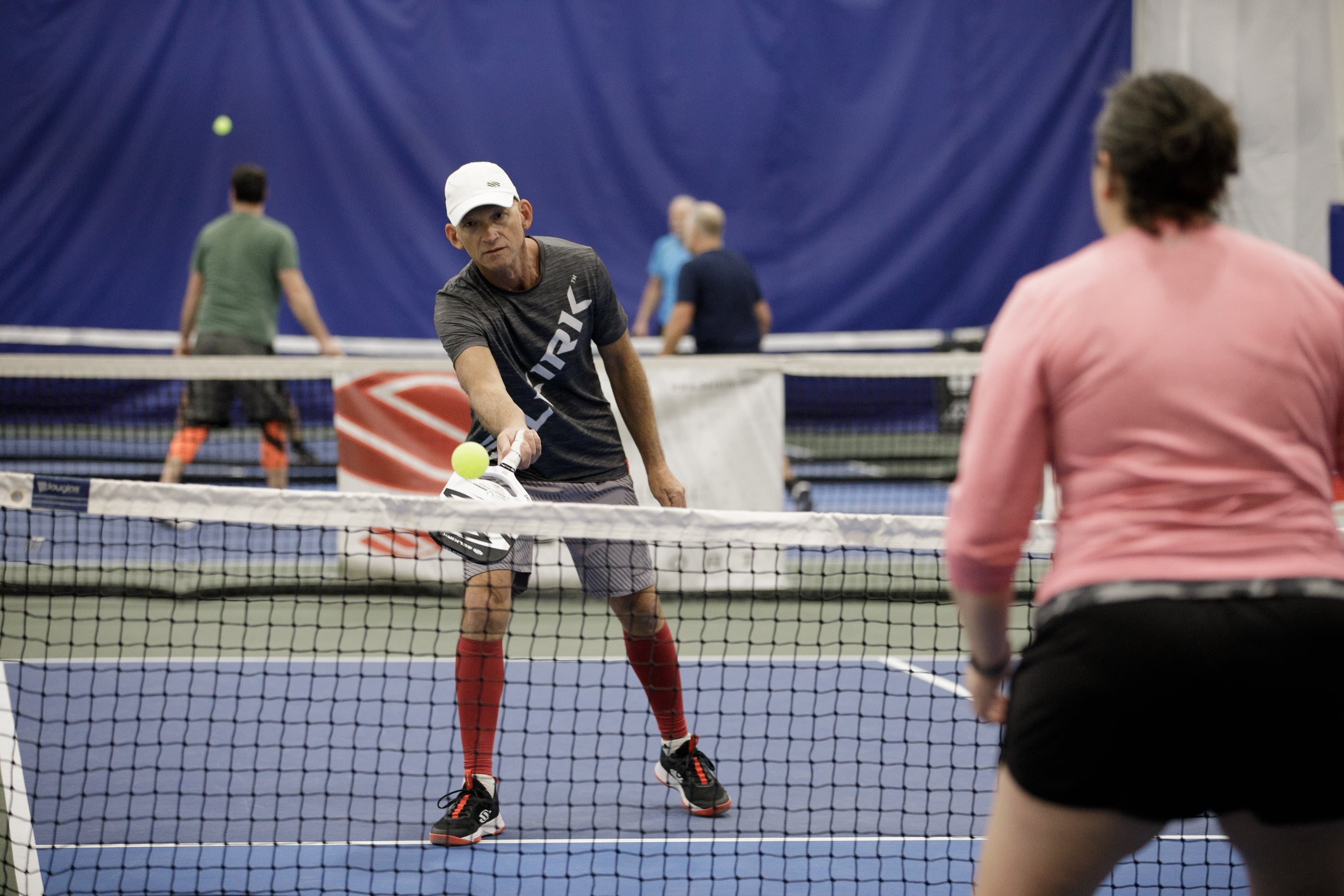 A player dinks a pickleball during a game, with other players in the background.