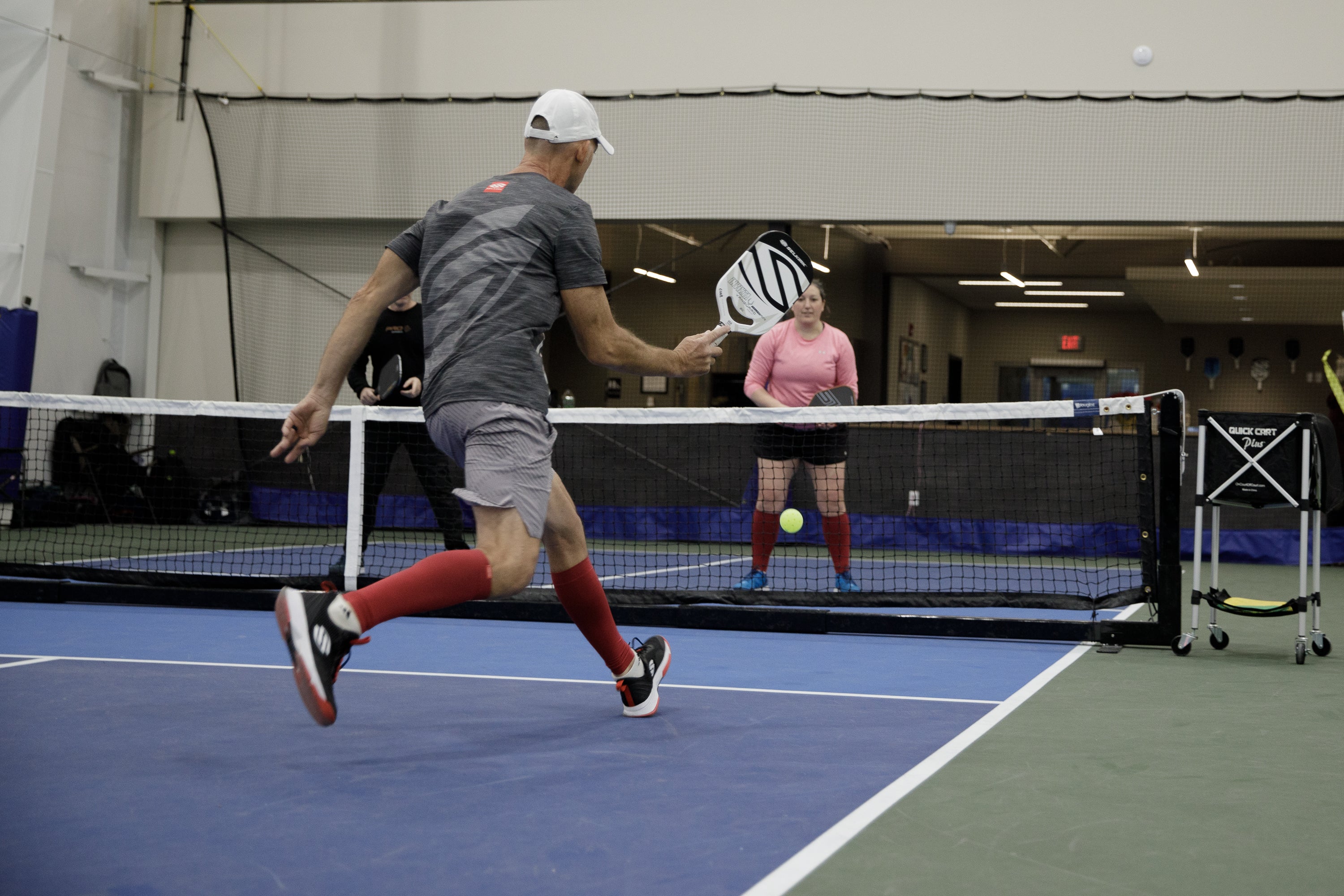 A player in a gray shirt and red socks dinks a pickleball over the net while another player prepares to return it.