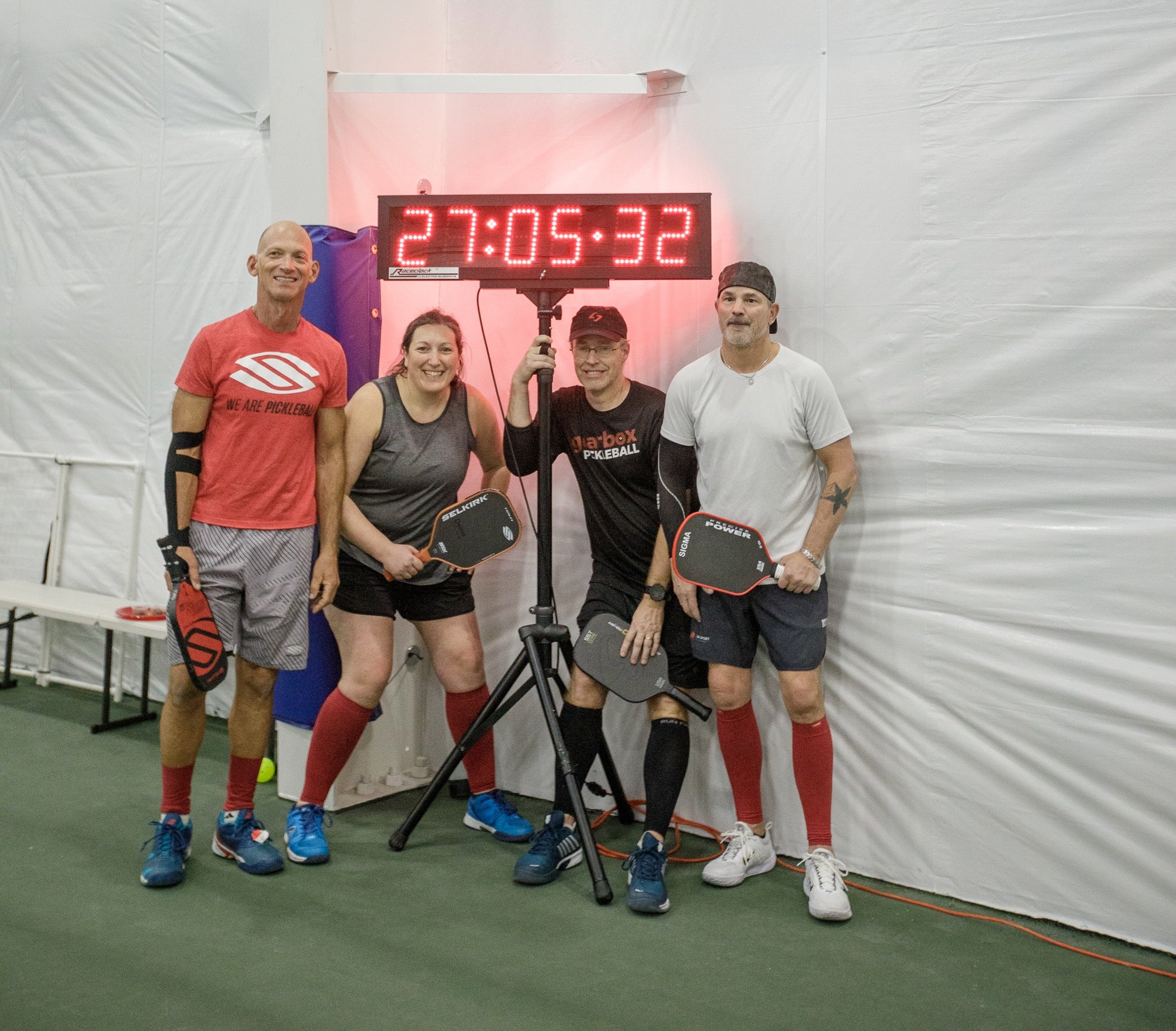 Four pickleball players pose in front of a timer showing 27:05:32 during a world record attempt.