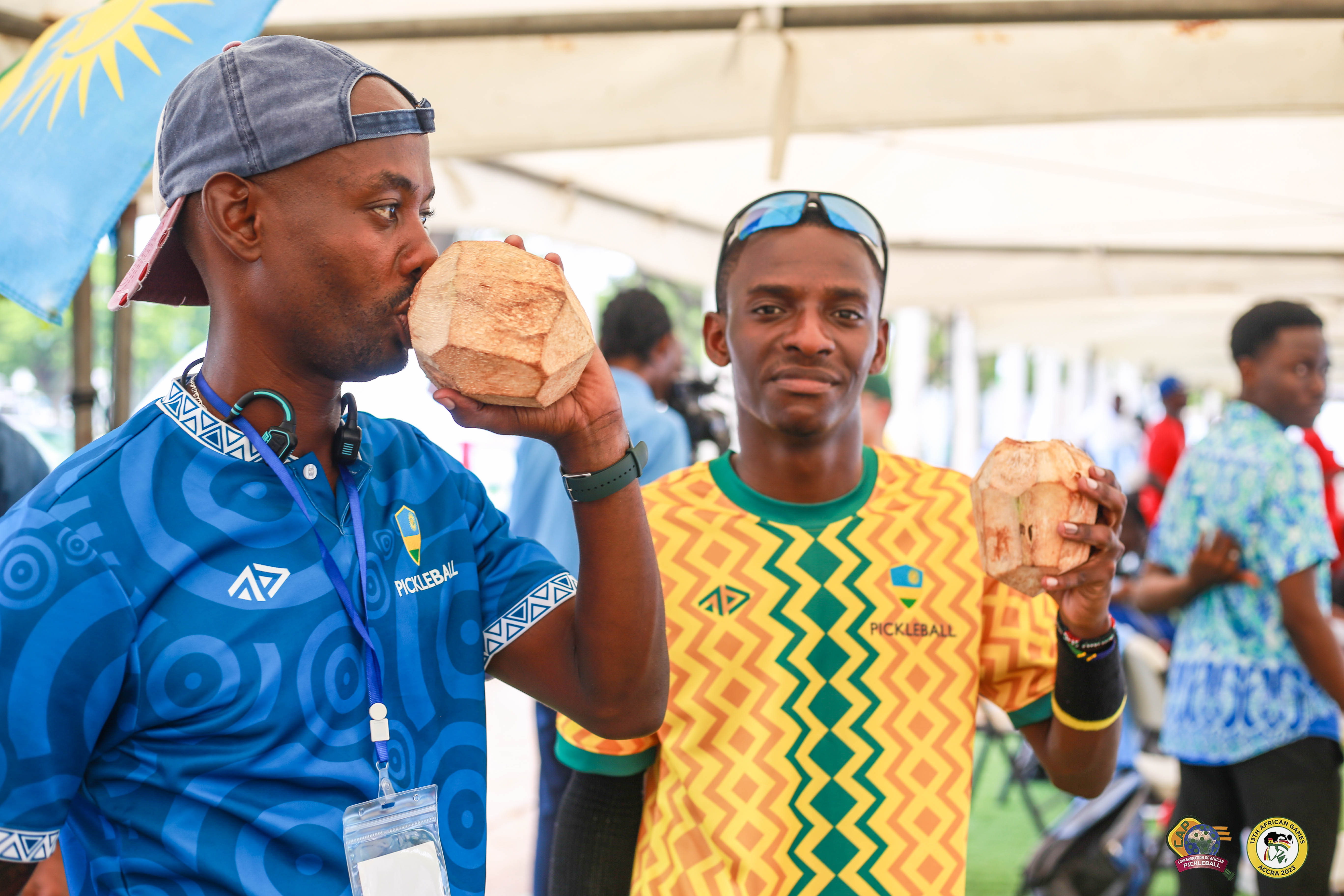 Two men holding coconuts, one in a blue shirt and the other in a patterned yellow shirt, at the African Games.