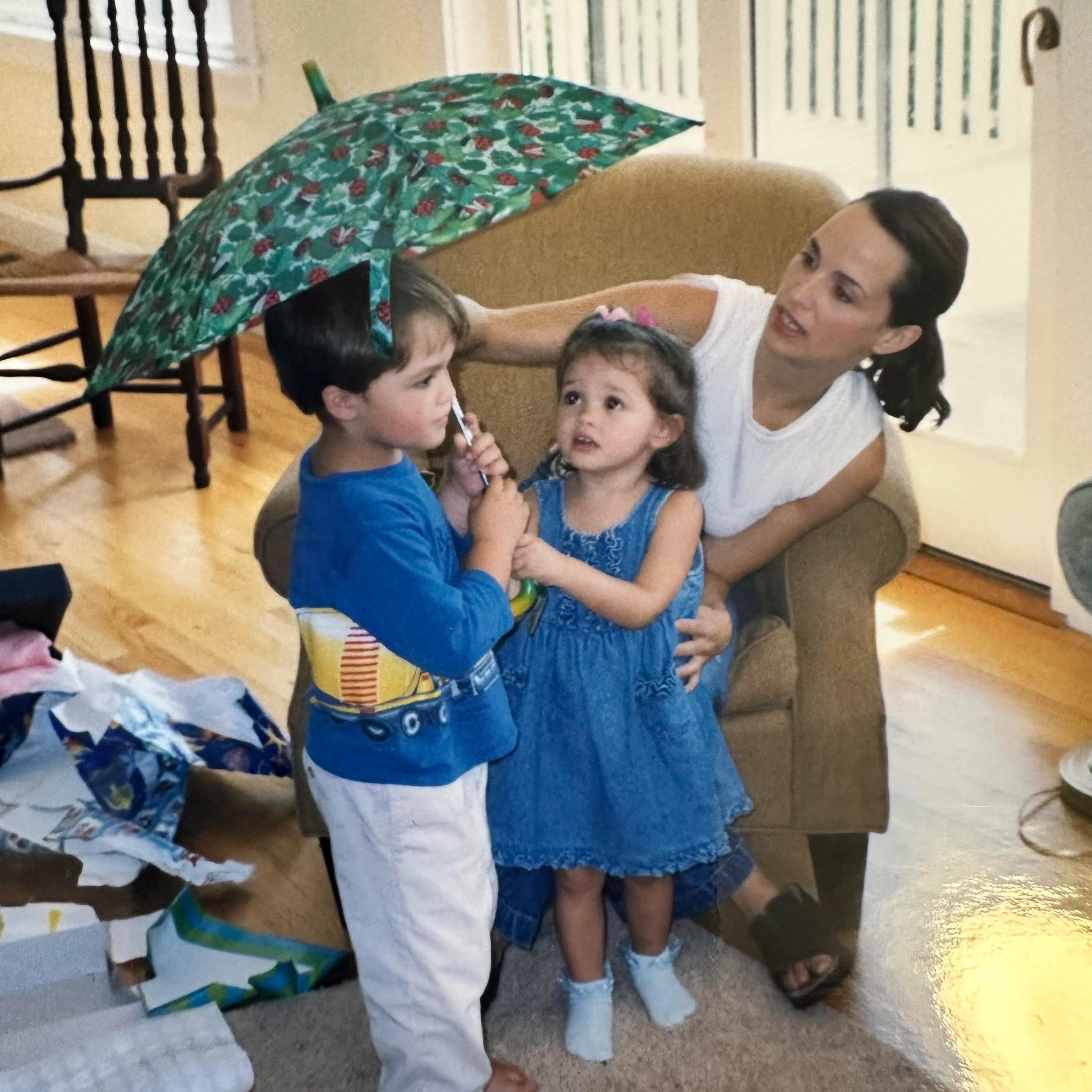 A mother sitting in a chair with her son holding an umbrella and daughter beside them, surrounded by wrapping paper.