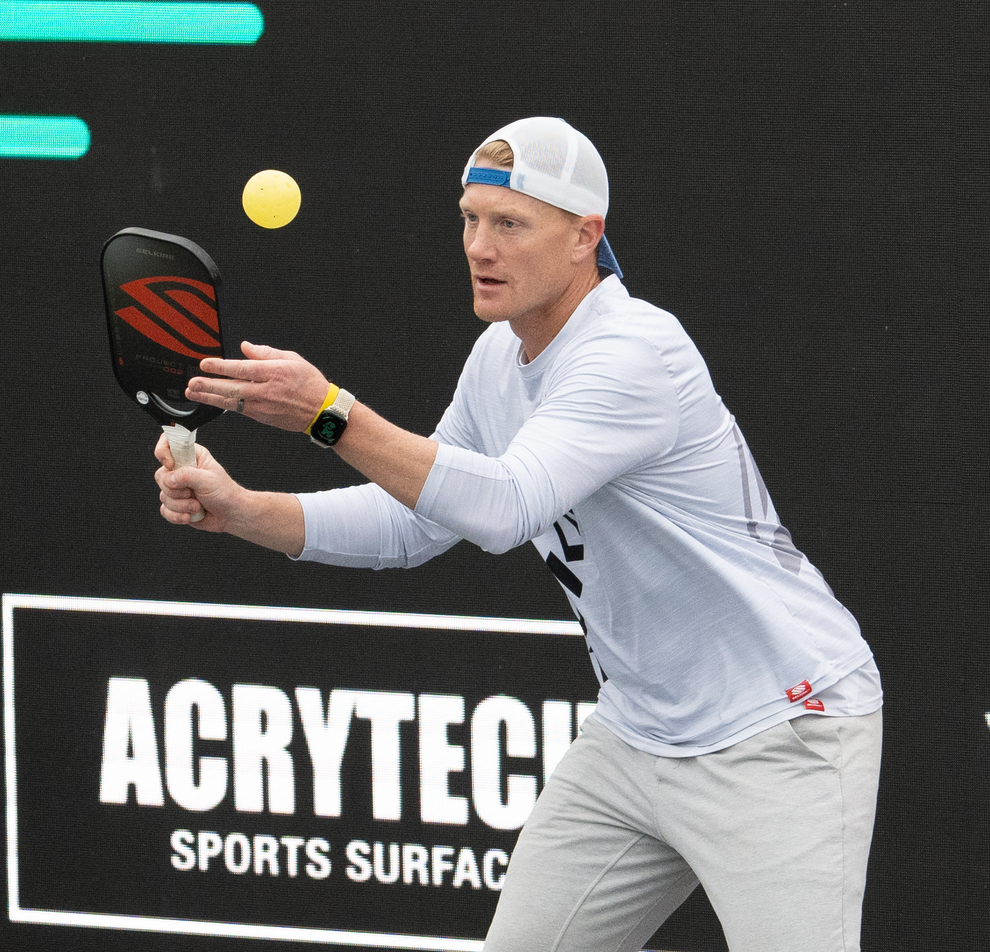John Cincola preparing to hit a pickleball during a match, wearing a white long-sleeve shirt and gray pants.