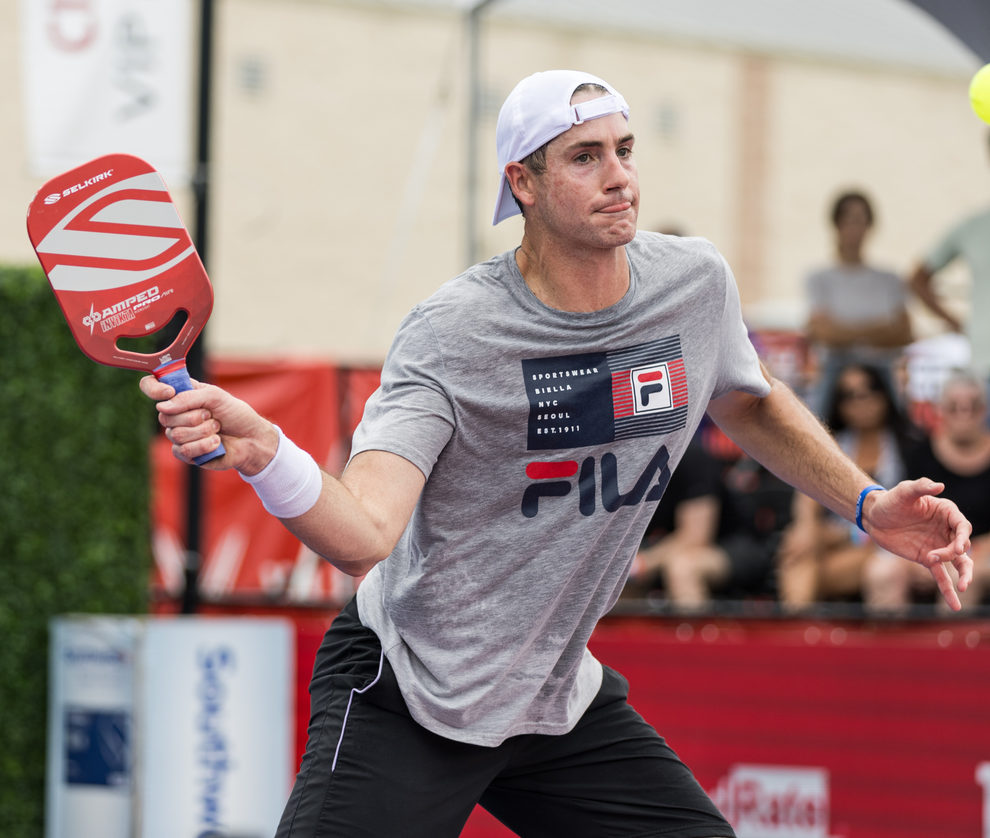 John Isner playing pickleball, holding a red paddle with 'AMPED' logo