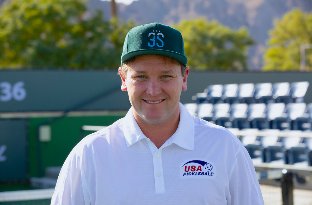 Smiling man wearing green cap 3S and white polo with USA PICKLEBALL logo at court, bleachers and 36 visible