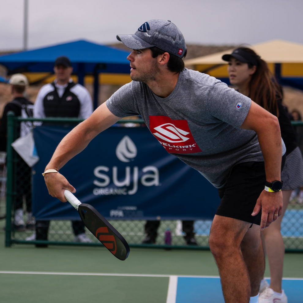 Male pickleball player in a gray cap and gray shirt with a red logo, preparing to hit a ball on an outdoor court with a 'suja organic' banner in the background.