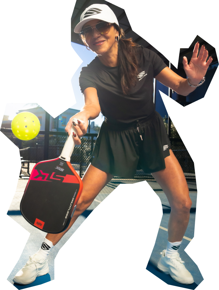 Woman playing pickleball with a paddle and ball, wearing a black outfit and white cap.
