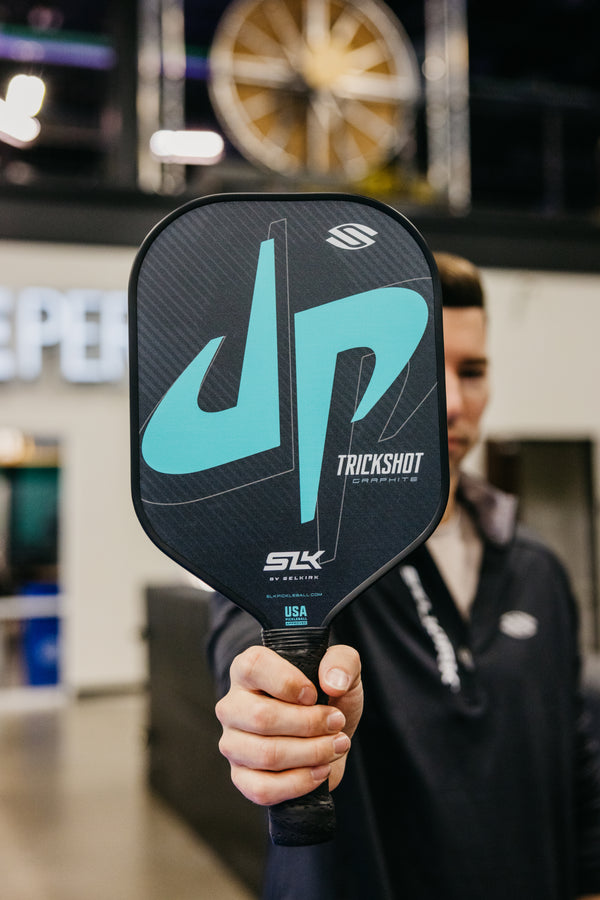 A person holding the SLK x Dude Perfect Trickshot pickleball paddle with a teal and black design.