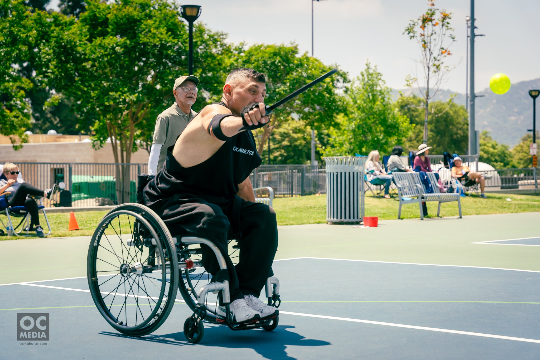 A person in a wheelchair playing pickleball on a court, with spectators in the background.