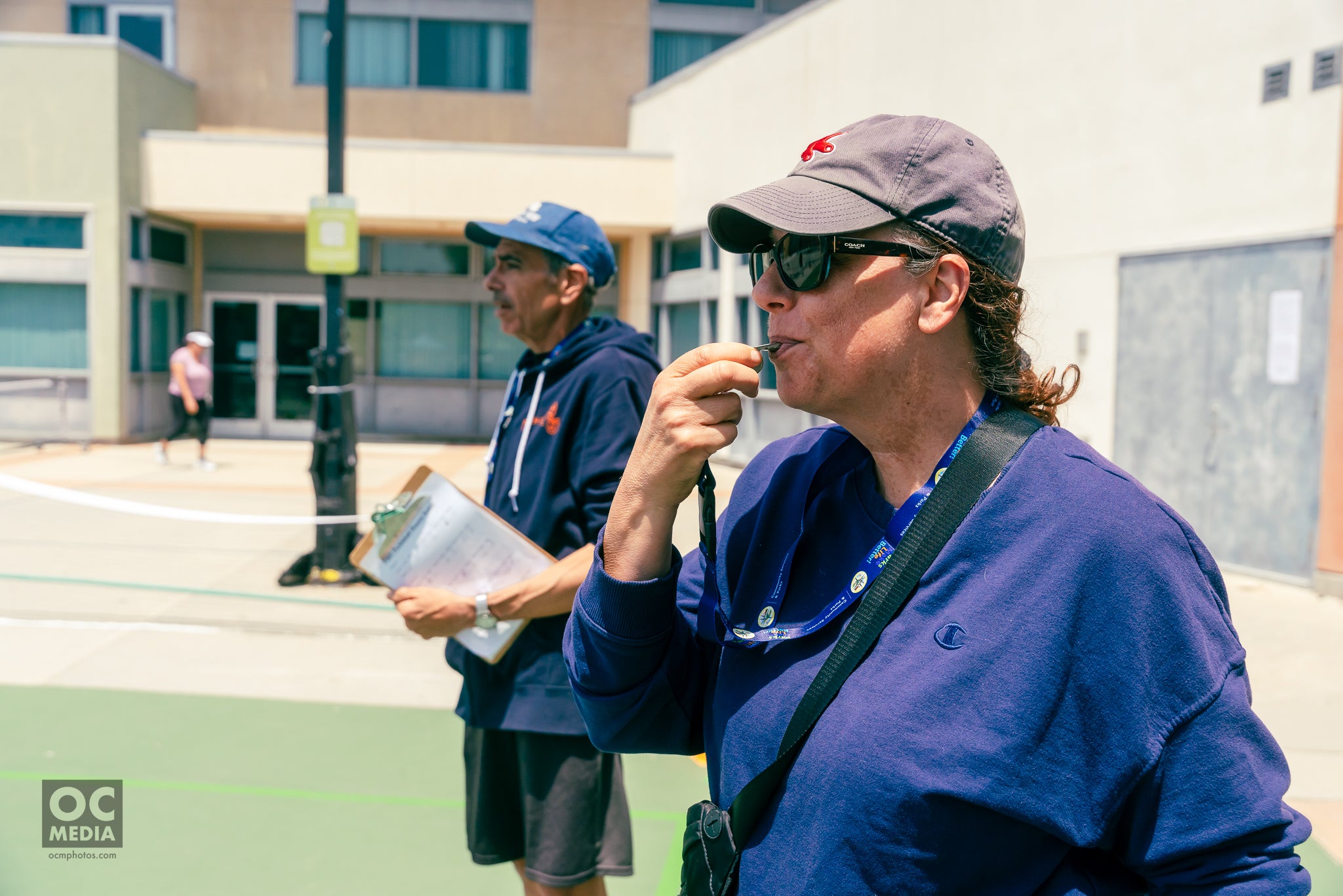 Two individuals overseeing a pickleball game, one holding a clipboard and the other blowing a whistle.