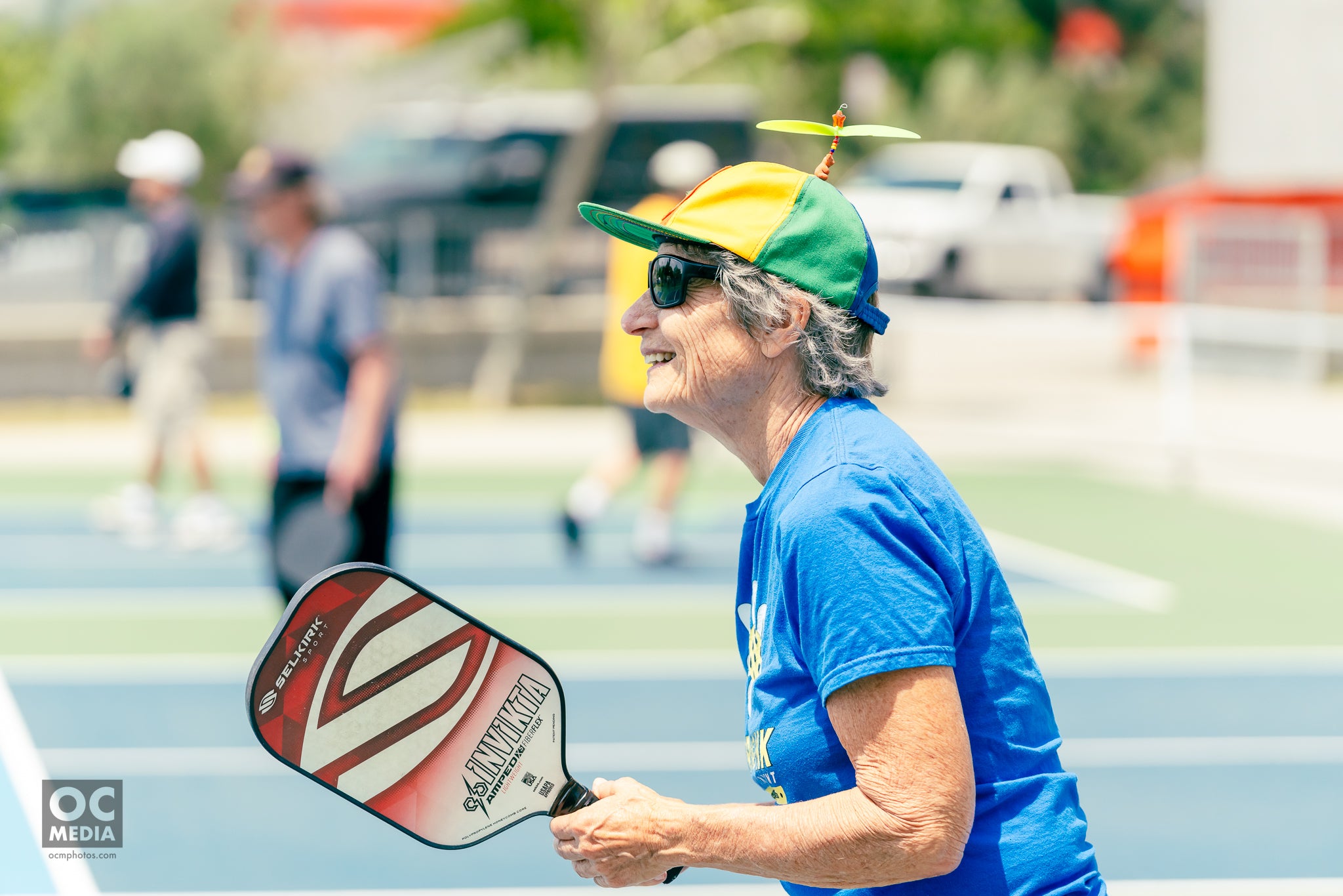 Older woman smiling while holding a pickleball paddle, wearing a colorful hat, at a pickleball event.