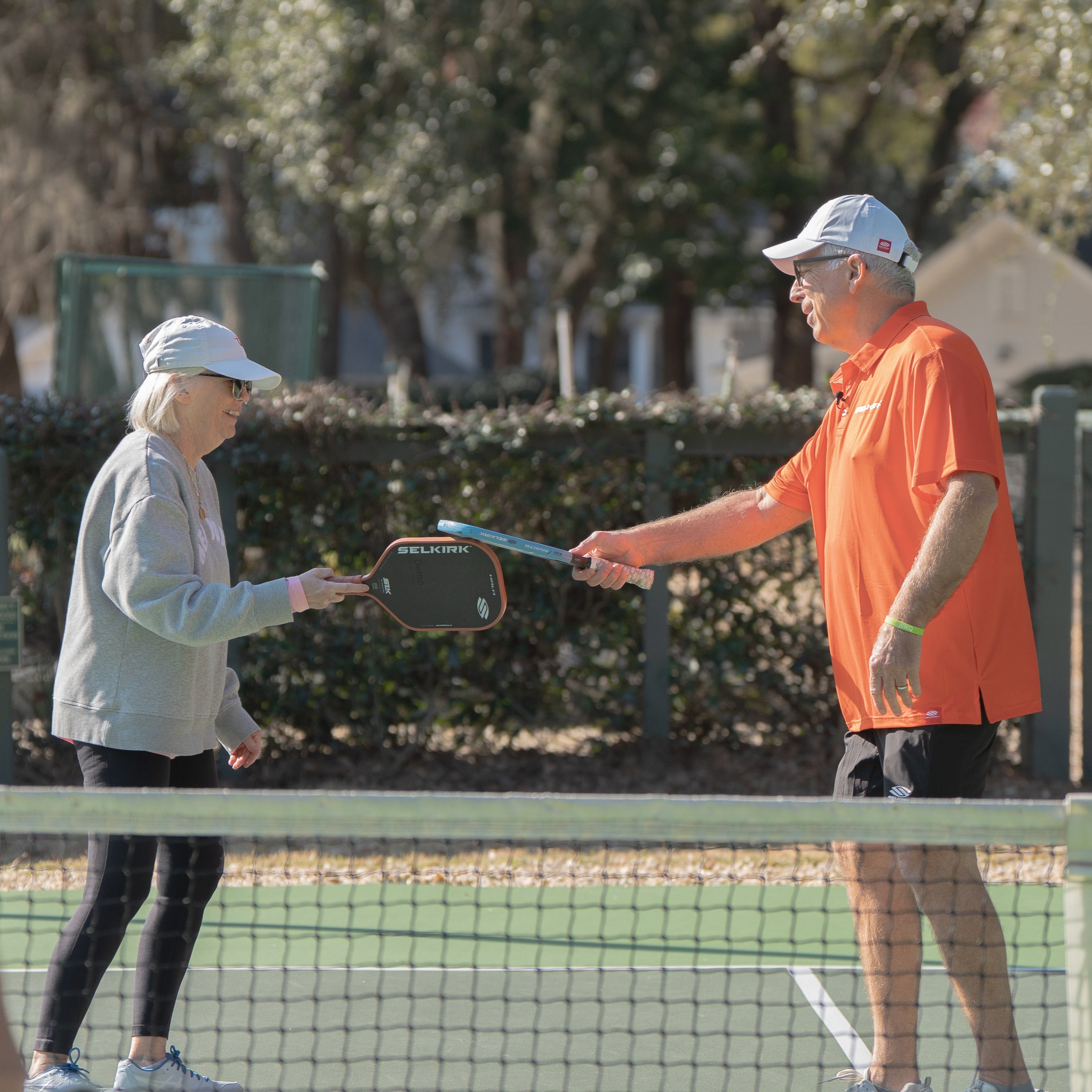 Two older adults on a pickleball court tap Selkirk paddles in a friendly gesture, with a third person in the foreground.