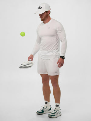 Male athlete in white shirt, shorts and cap holding a pickleball paddle while watching a green pickleball in midair.