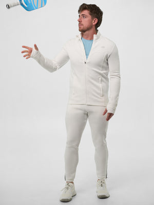 Man wearing the Pro Line Full Zip Jacket in Chalk and matching joggers tossing a blue pickleball paddle in a full-length studio shot.