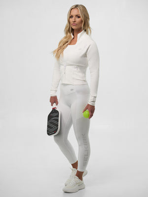 Woman in white athletic jacket and leggings holding a pickleball paddle and neon green pickleball; leggings read SELKIRK.