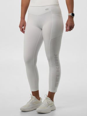 Women's white high‑waist 7/8 leggings with perforated 'SELKIRK' logo on lower right leg, shown with white athletic sneakers.