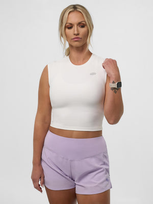 Woman wearing white sleeveless cropped top and lavender high-waist athletic shorts, standing with hand raised to wristwatch.