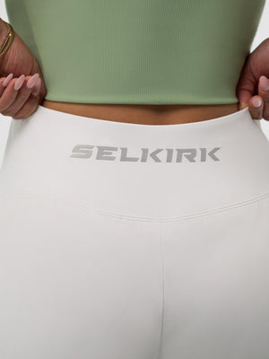 SELKIRK logo on back waistband of white high-rise leggings; hands adjusting waistband, cropped torso visible