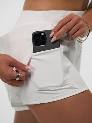 Hands placing a smartphone into a side pocket of a white athletic skort, showing pocket size and seam details