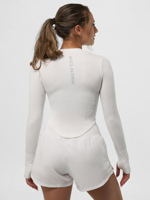 Woman seen from back wearing white long-sleeve athletic top labeled SELKIRK and white running shorts, hair in a ponytail.