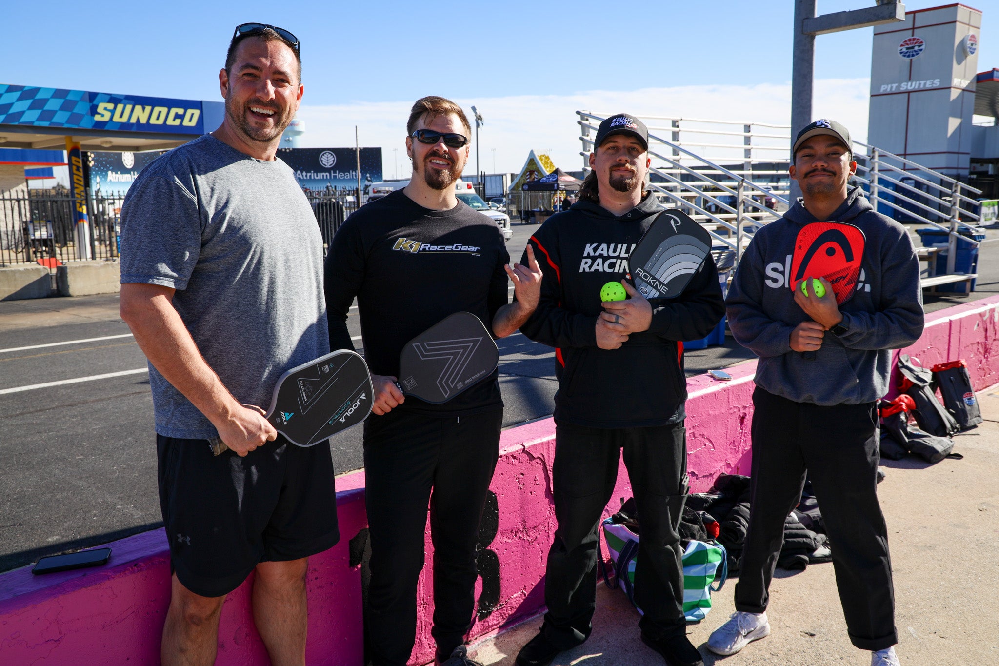 Four NASCAR drivers holding pickleball paddles and balls, smiling outdoors.
