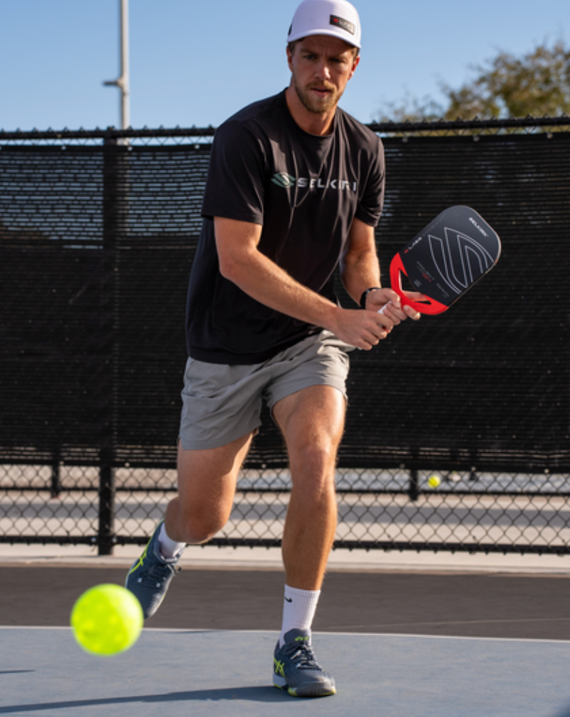 Man wearing a SELKIRK shirt and cap lunging on a court to hit a neon green pickleball with a paddle.
