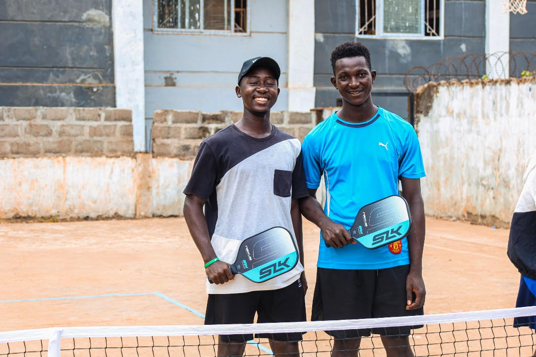 Two boys smile at the camera as they hold up two SLK Nexus paddles. They stand on a dirt pickleball court. 
