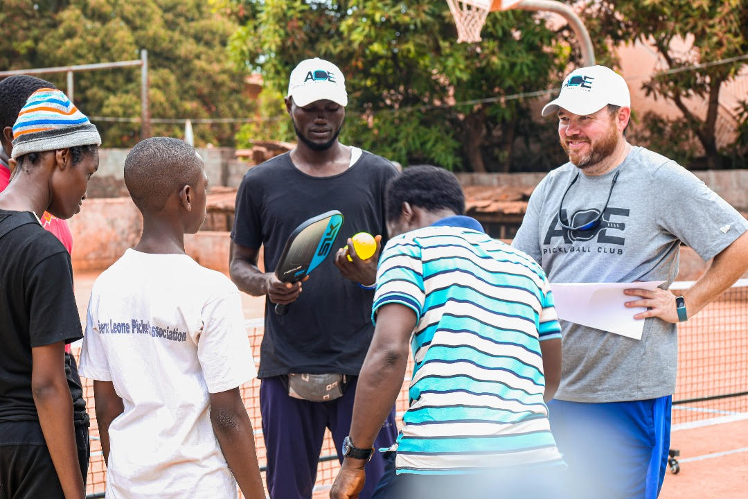 Children gather around two men who are explaining rules. They stand on a dirt pickleball court near the net. 