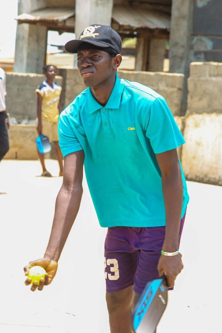 A child stands at the baseline of a dirt pickleball court. He holds a pickleball in front of him as he prepares to serve. 