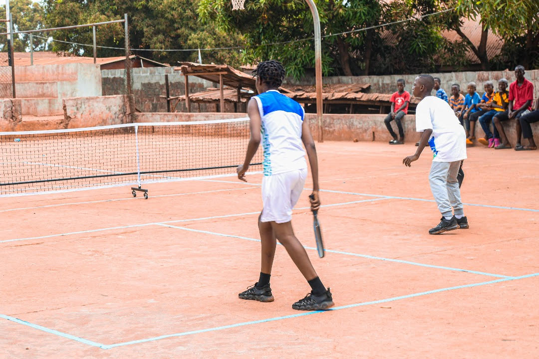 Two children stand on a dirt pickleball court near the baseline. They are both waiting to receive the next shot. A crowd of children watches the game. 