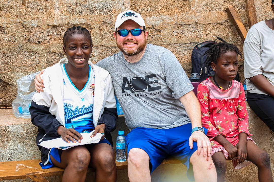 A boy and two girls sitting together, smiling, at a pickleball event in Sierra Leone.