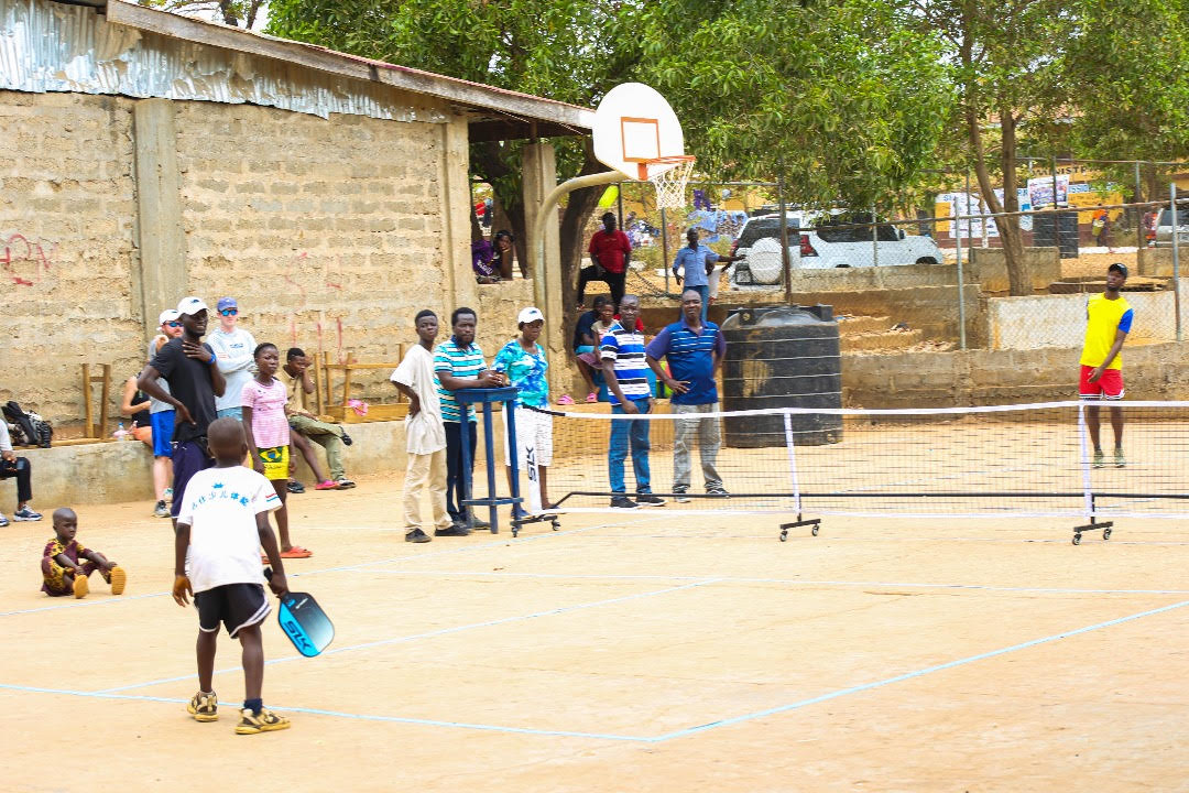 A small child stands at the baseline of a dirt pickleball court. He is ready to receive a serve. Adults stand nearby to help explain the rules. 