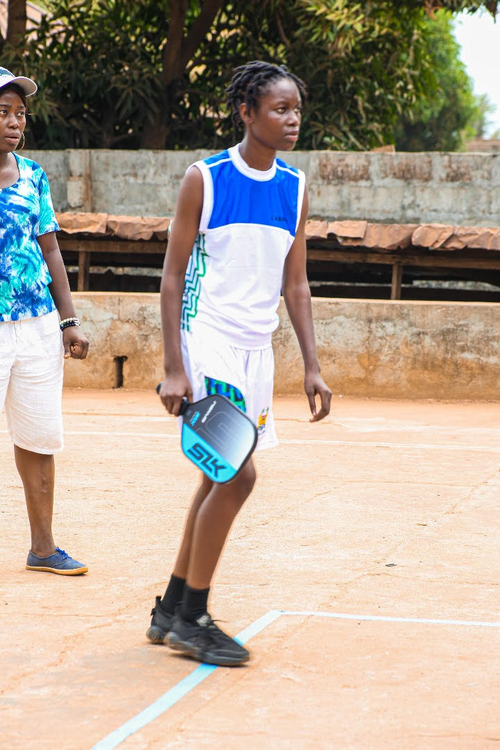 A student holds their Selkirk Nexus paddle while standing at the baseline of a dirt pickleball court. They are preparing to receive a serve while an adult gives advice off to the side. 