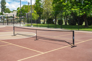 SLK Pro Portable Pickleball Net set up on a court with a brown surface and green trees in the background.