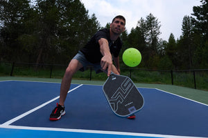 A player in a black shirt and shorts reaching for a green pickleball with an SLK HALO Control paddle on a blue court.