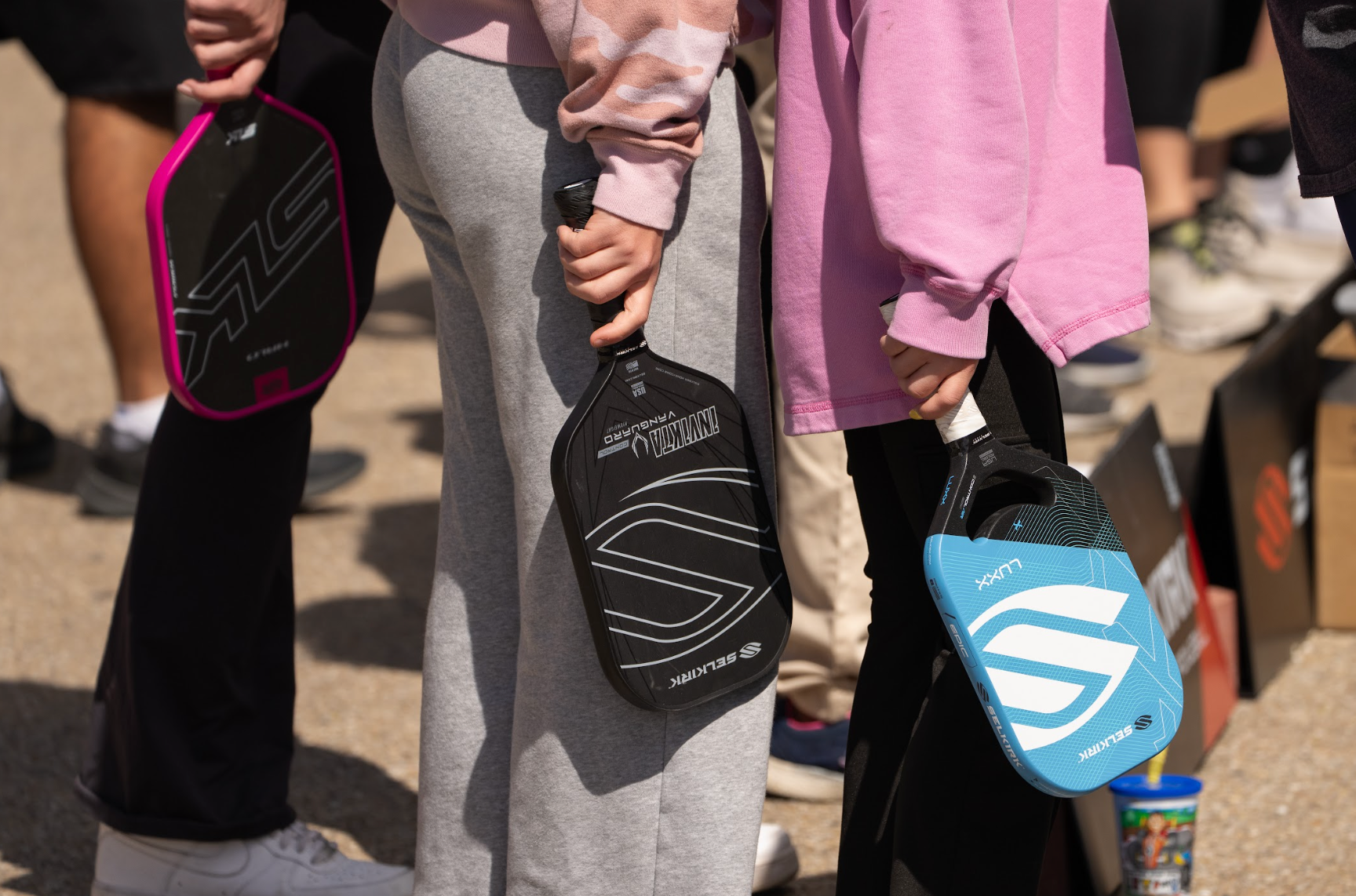 Three children hold Selkirk pickleball paddles next to an outdoor court. 