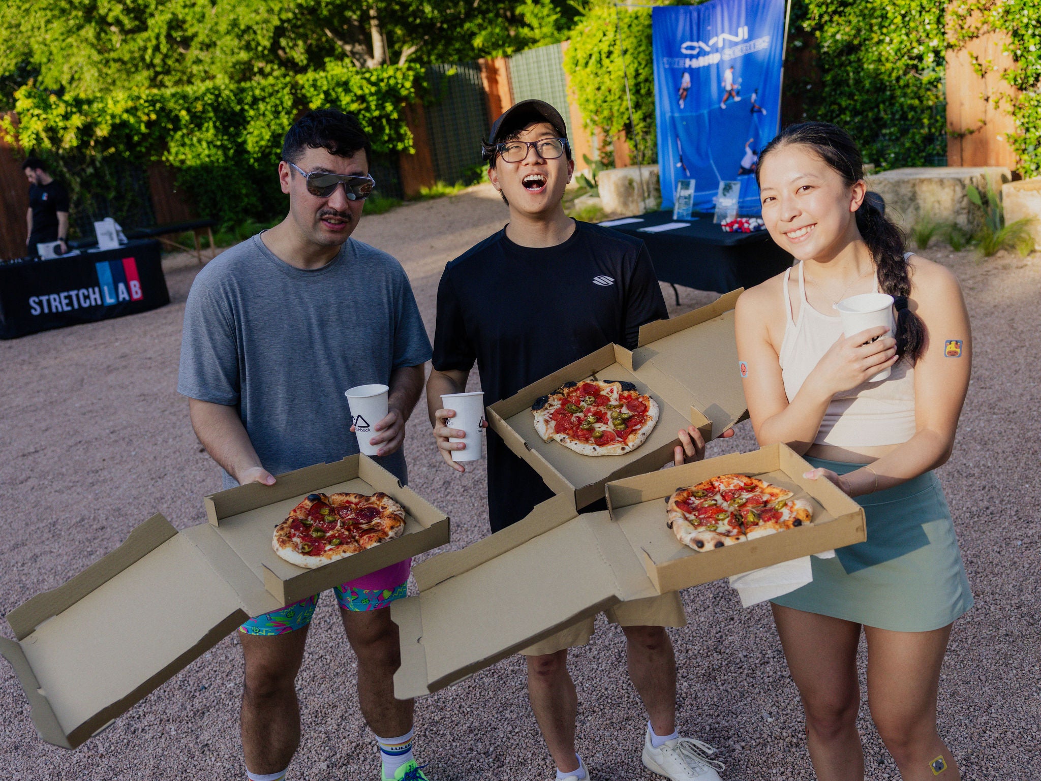 Three people smile as they eat pizza from to-go pizza boxes. 
