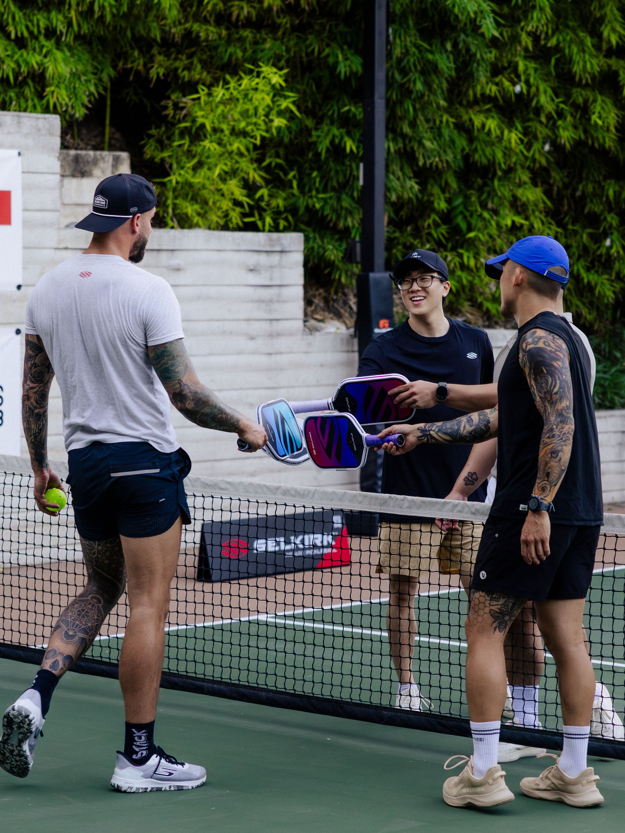 Four men tap pickleball paddles over an outdoor net. 