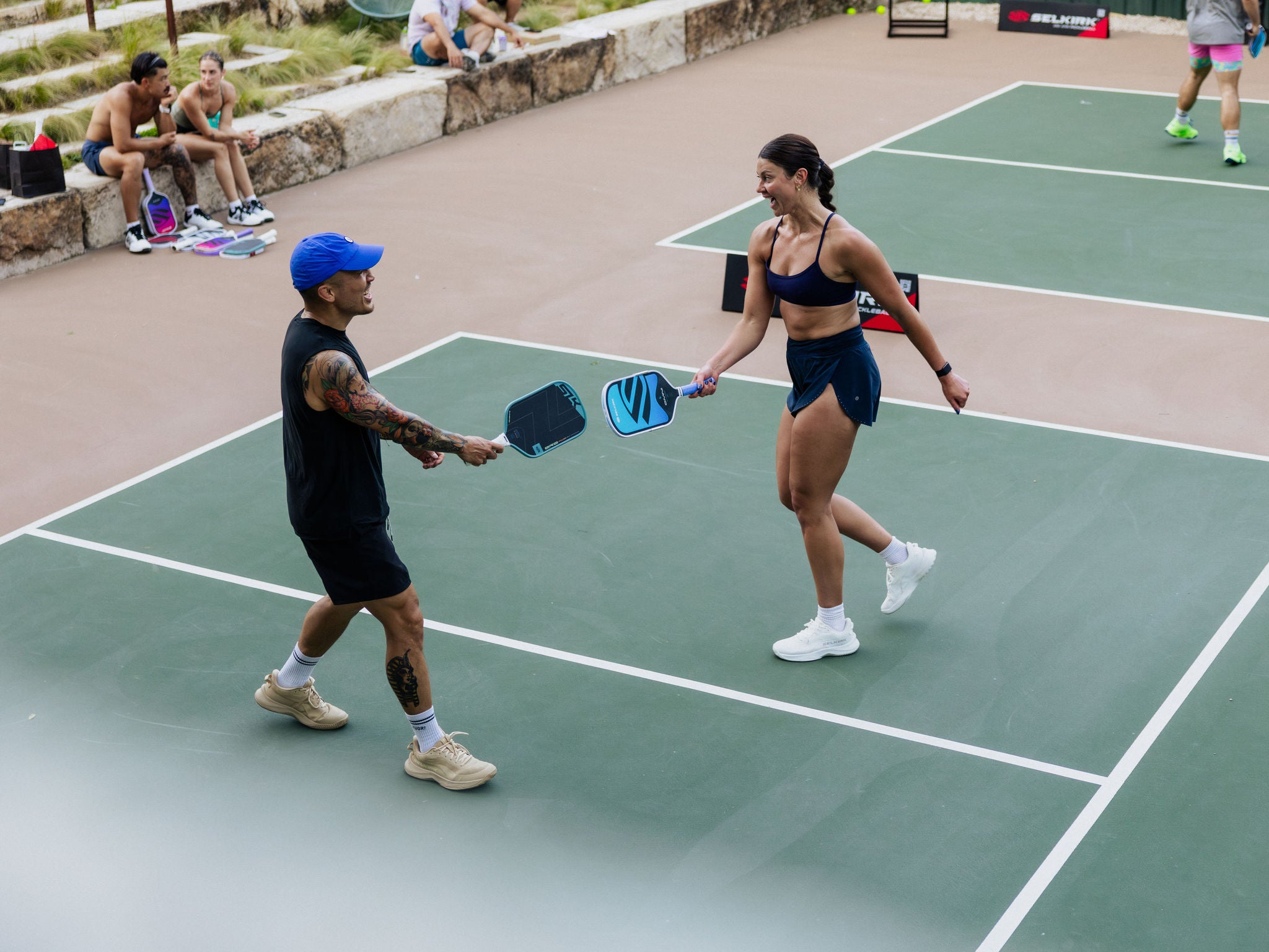 A man and a woman smile and tap pickleball paddles on an outdoor court. 