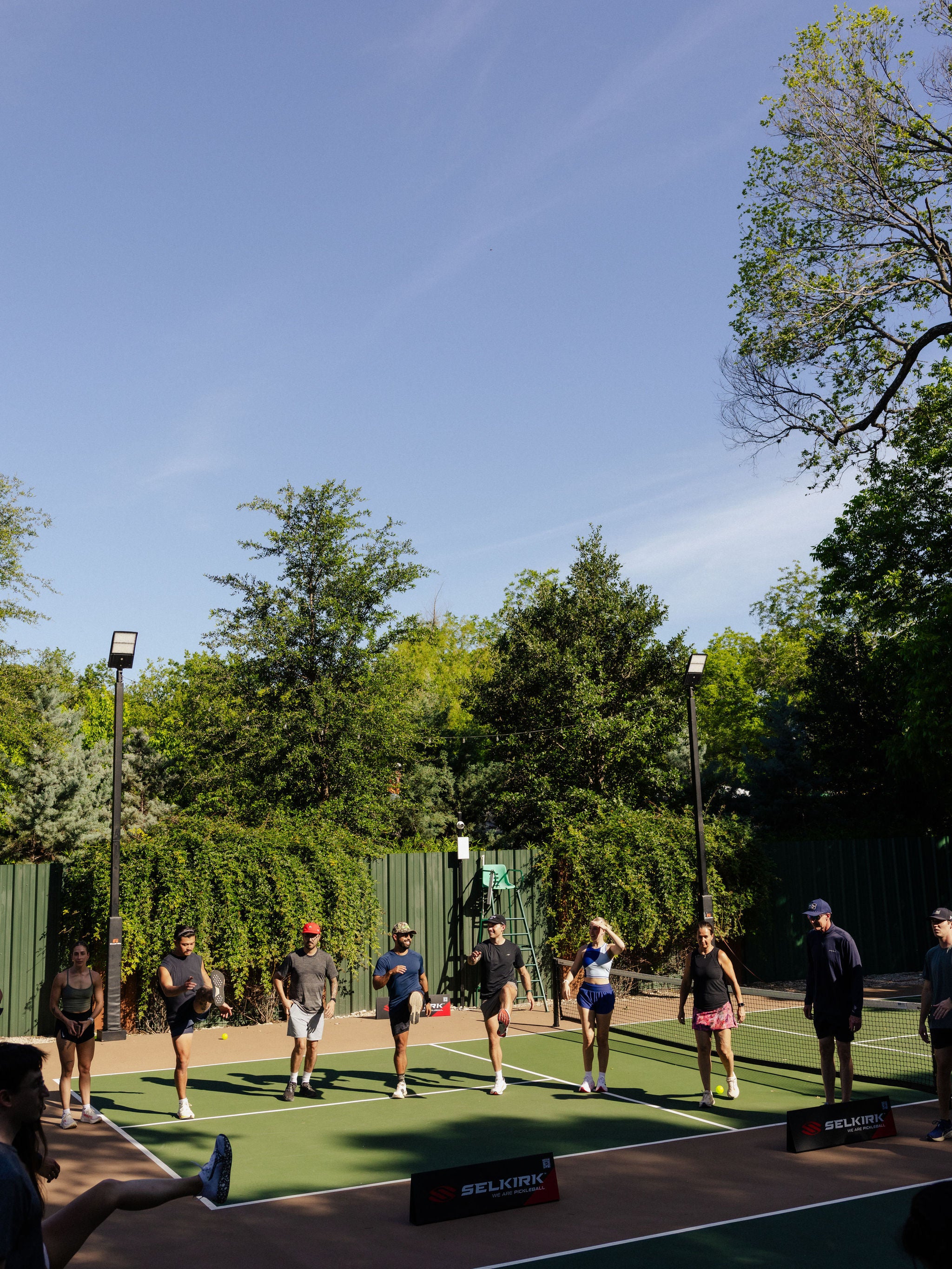 A large group stretches in a circle on an outdoor pickleball court. 