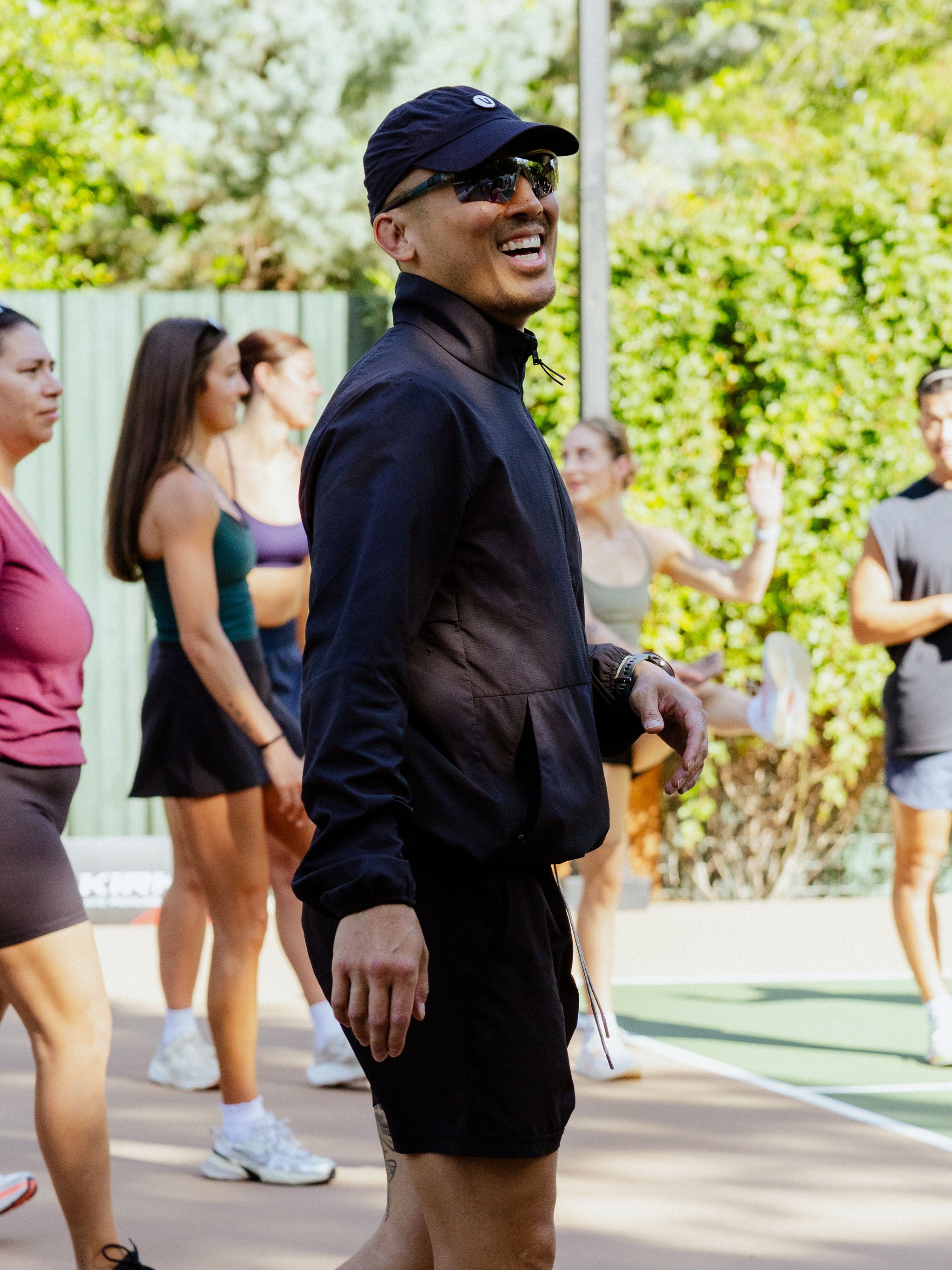 A man smiles as he stands in a group of people stretching on an outdoor pickleball court. 